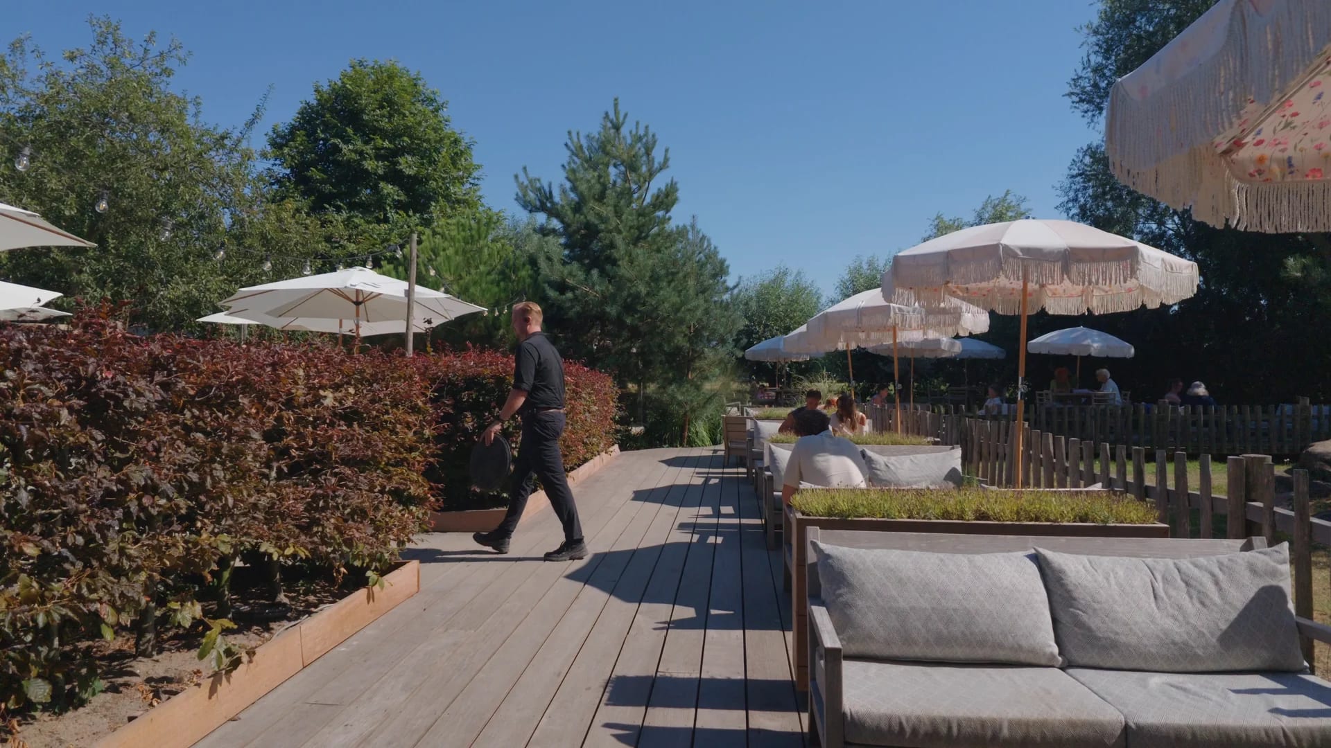 Een medewerker in het zwart loopt over de houten vlonder langs de rode beukenhaag van het loungeterras, parasols en gasten in de zon verderop.