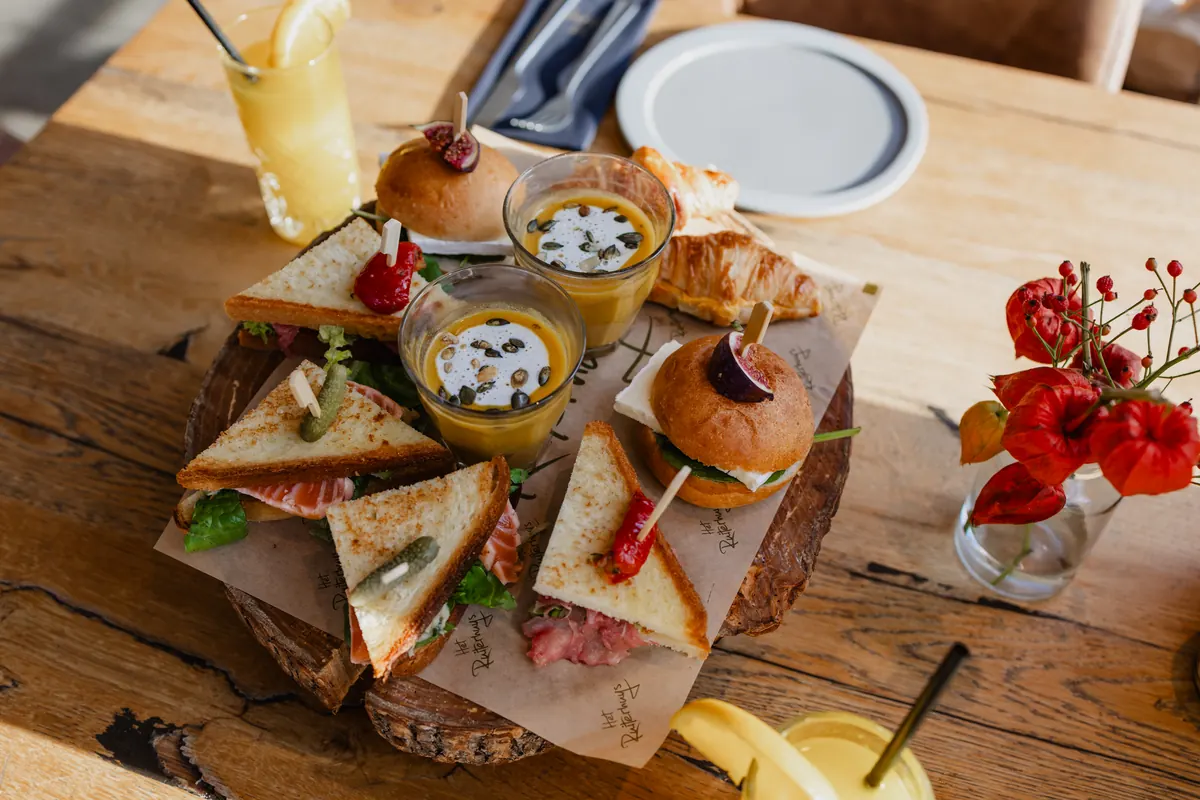 Een boomschijf met bakpapier waarin het Ruiterhuys-logo is gedrukt, vol geroosterde sandwichpunten met zalm en sla, een punt met tartaar en een rode peper op een prikker, twee zachte broodjes waarvan één met een halve vijg, en een croissant. Ertussen twee glaasjes oranje soep met een dot room en pompoenpitten. Rechts een glazen vaasje met rode lampionnetjes, linksboven een hoog glas jus d’orange met een schijf citroen, en achteraan een leeg grijs bord met bestek. Zonlicht ligt in strepen over het ruwe eiken tafelblad.