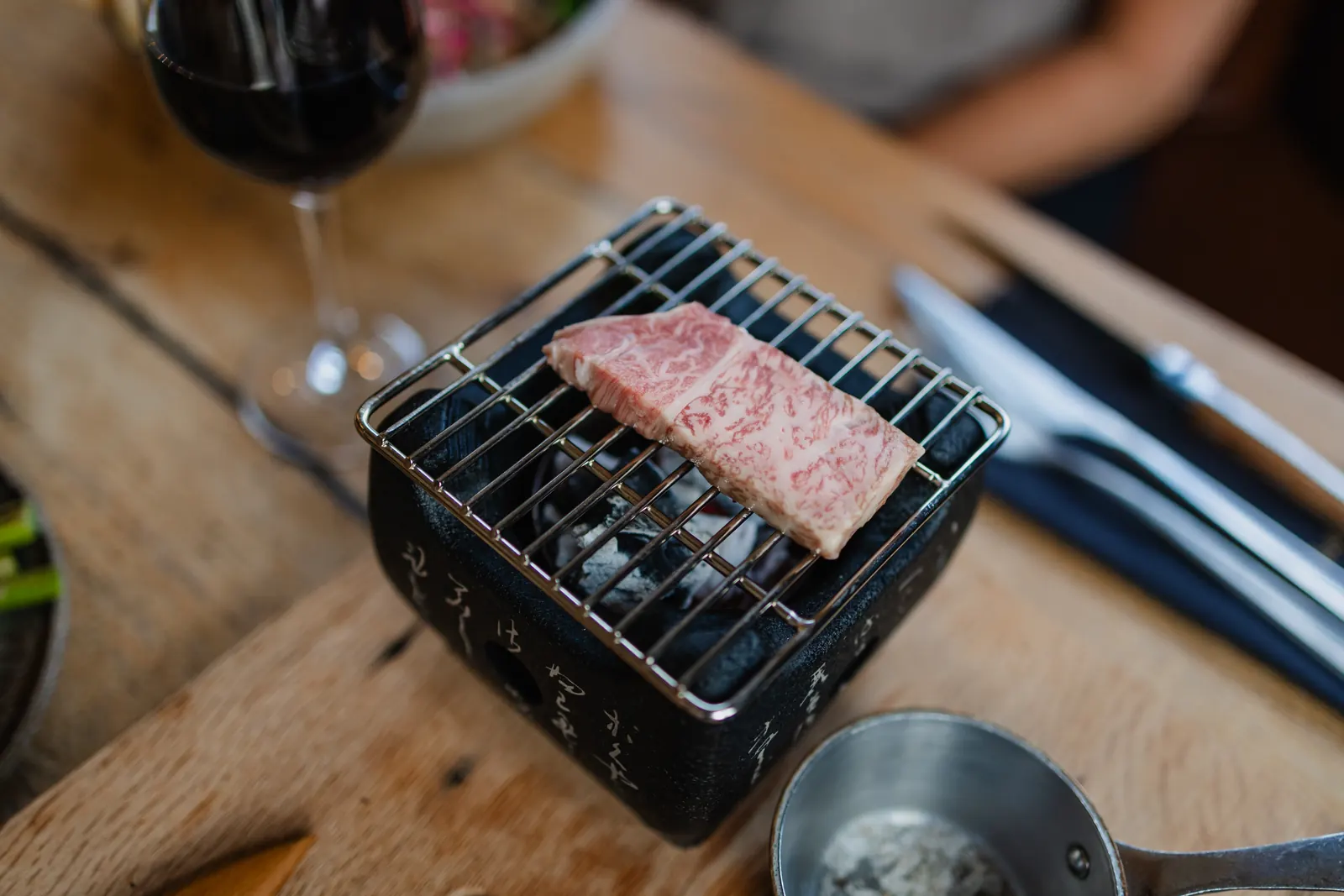 Een klein zwart grilltafeltje met Japanse tekens op de zijkant staat midden op een eiken tafel; onder het stalen rooster gloeien de kolen. Bovenop ligt één dikke plak rundvlees, diep gemarmerd en nog rauw. Links op de voorgrond, onscherp, een glas rode wijn; rechts mes en vork, en rechtsonder een stalen steelpannetje met saus.