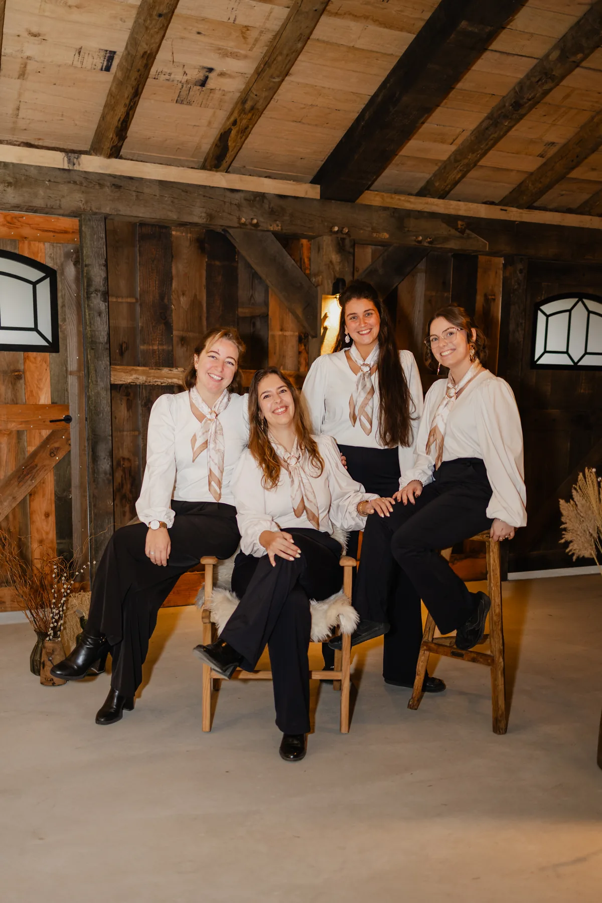 Vier collega's van de bediening bij elkaar in de oude stal, in witte blouses met een beige sjaaltje en een zwarte broek: twee zittend op houten krukken met een schapenvacht, twee erachter, allemaal lachend naar de camera. Achter hen de verweerde houten wand met twee glas-in-loodraampjes en een bos droogbloemen.