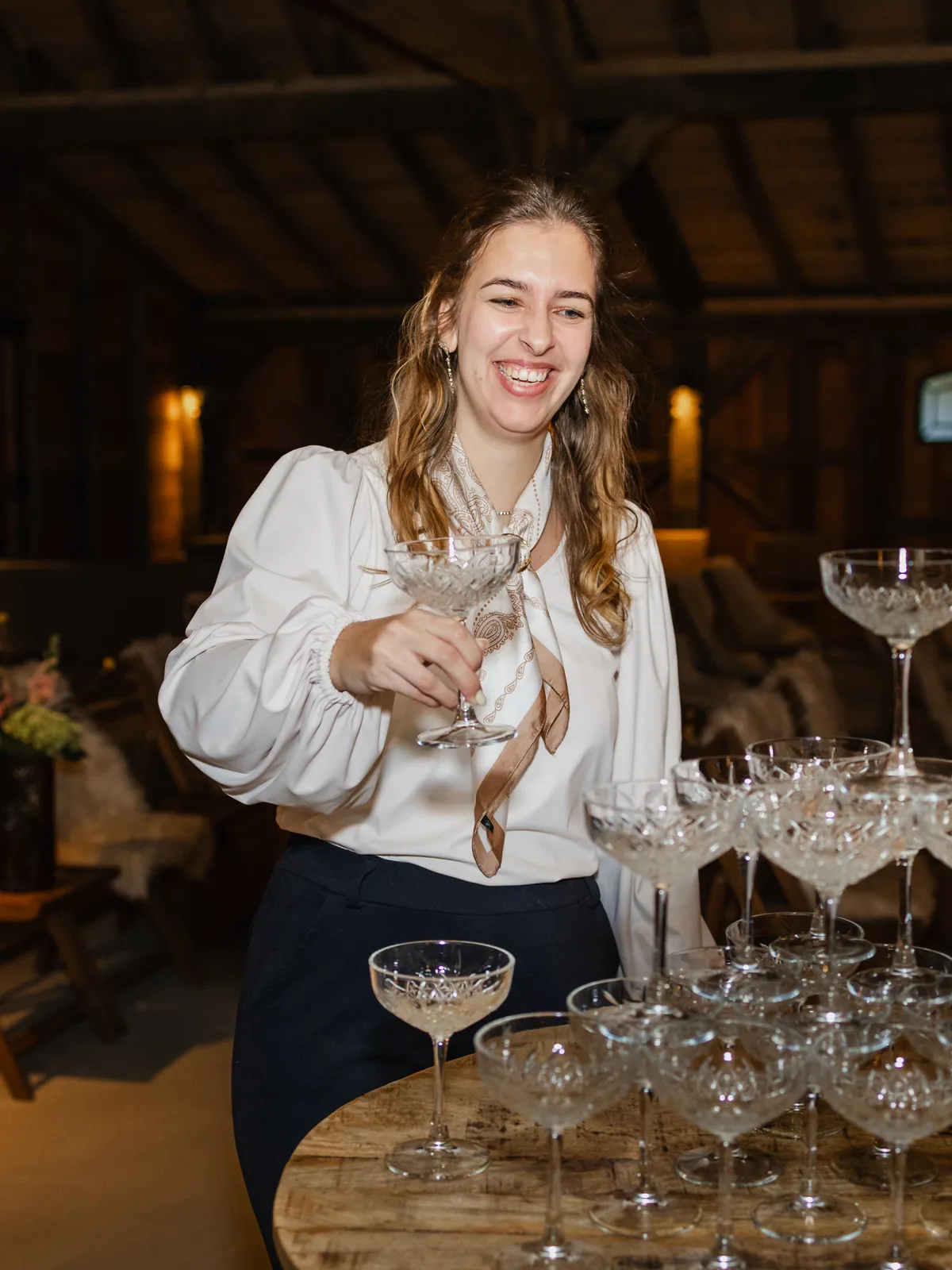 Sanne staat lachend achter een toren van champagnecoupes met een glas in haar hand, in een witte blouse met pofmouwen, met de stoelen en de kap van de schuur achter haar.