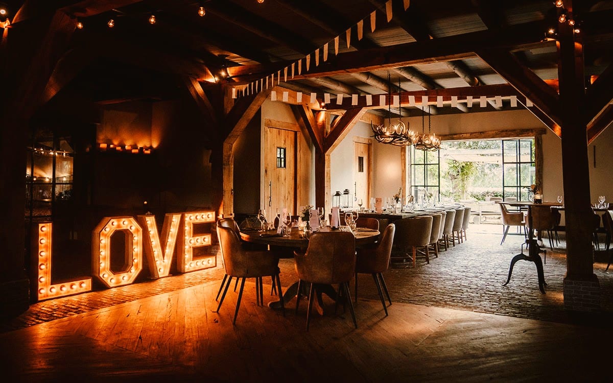 De Grote Zaal aangekleed voor een feest: witte vlaggetjes en lichtsnoeren tussen de donkere houten balken, twee geweikroonluchters boven een lange gedekte tafel, op de voorgrond een ronde tafel met leren stoelen en links verlichte LOVE-letters op de houten vloer. Achterin stalen puien met zicht op het groen.
